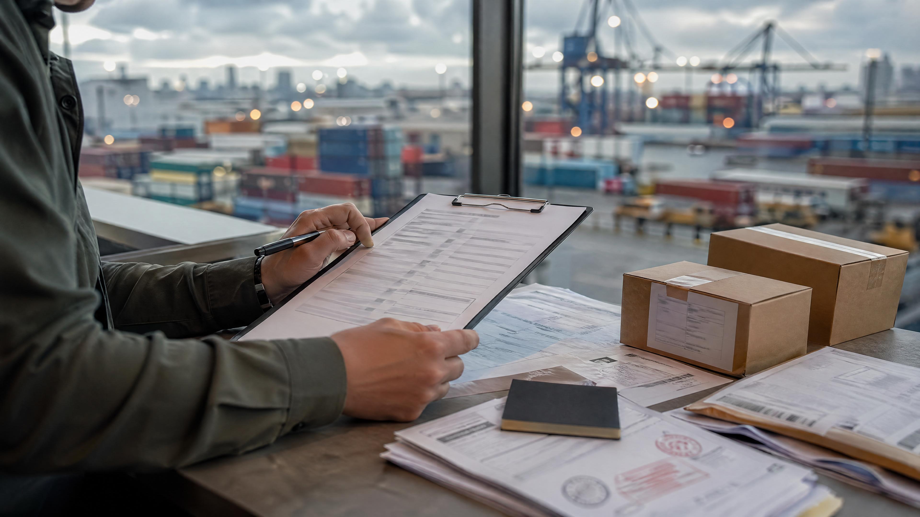 Editorial photo of customs paperwork, product cartons, a broker contact card, and blurred port logistics context for Shopify importing.