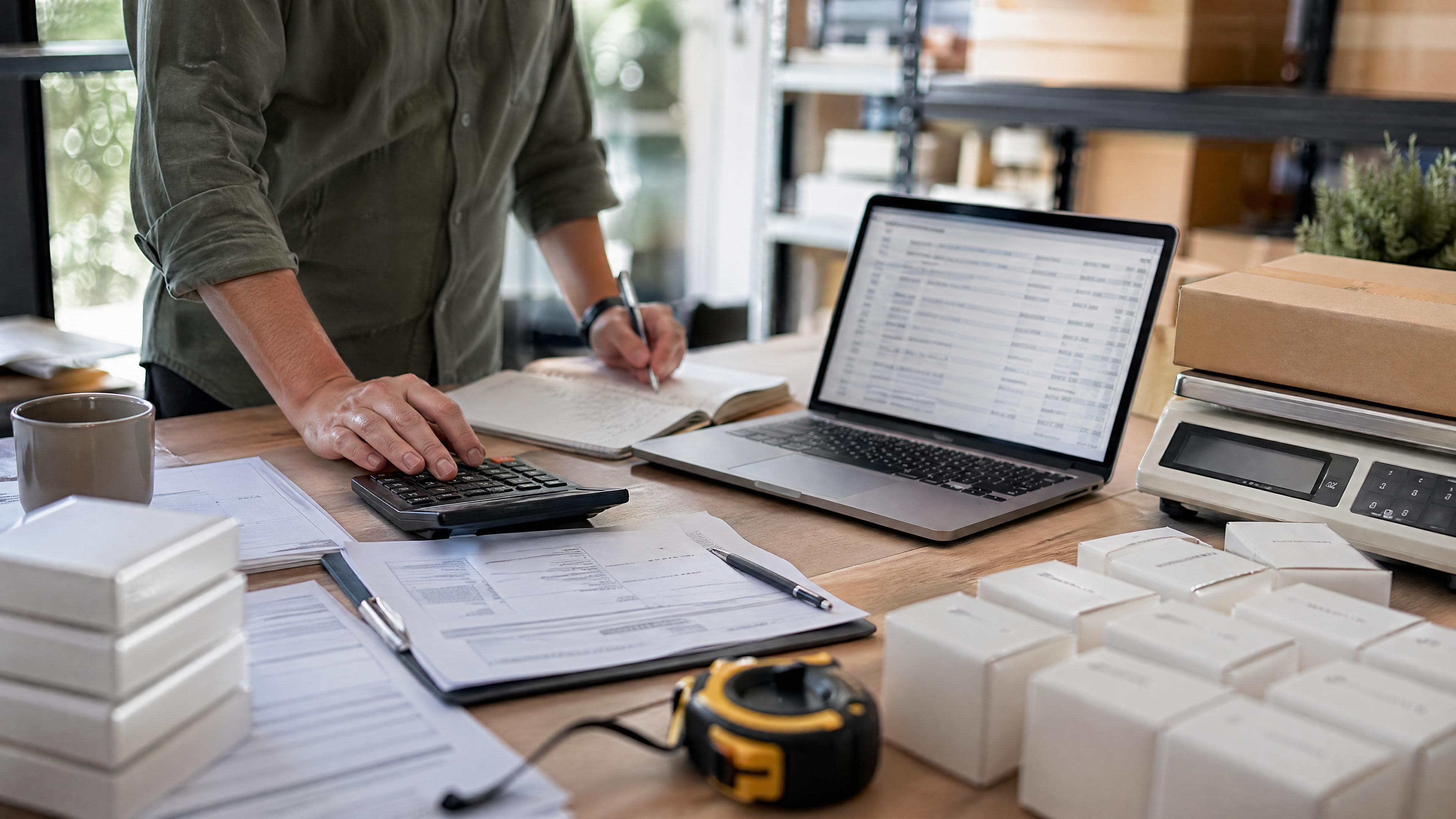Editorial photo of an ecommerce importer table with product units, freight invoice, customs forms, measuring tape, scale, calculator, and blurred landed-cost spreadsheet.