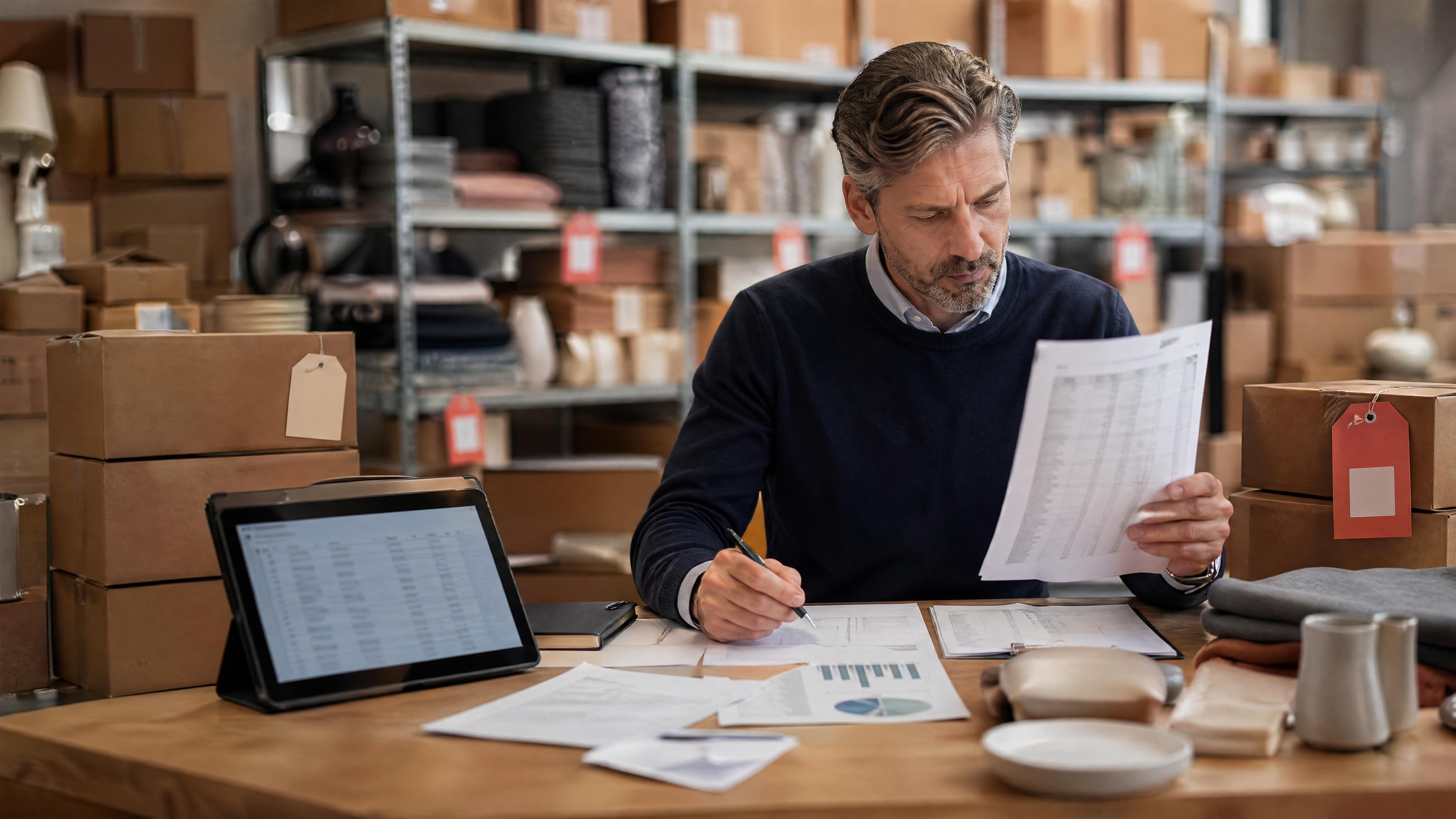Editorial photo of unsold retail inventory, boxed home goods, markdown planning, and a blurred inventory tablet in a stockroom.